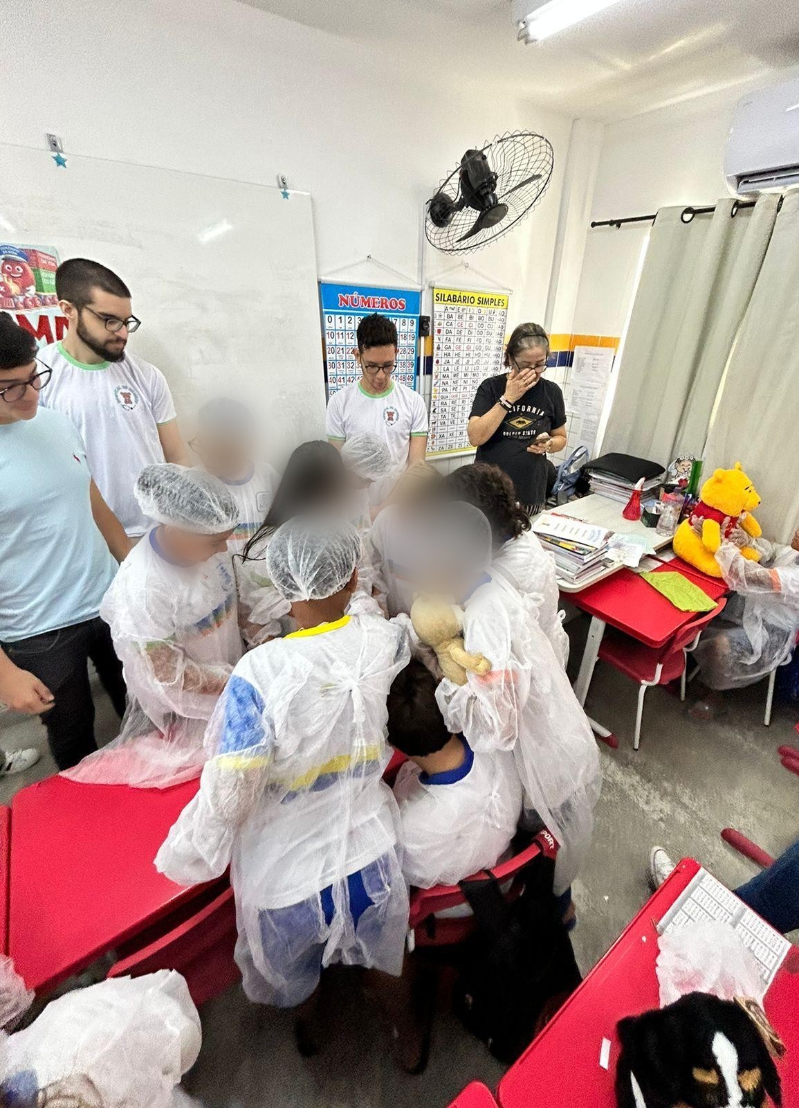 Crianças de jaleco e touca reunidas em torno de um ursinho de pelúcia durante uma estação, com integrantes ao lado